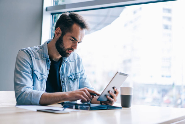 Focused bearded entrepreneur in casual clothes sitting at table with documents and browsing tablet while working on project in contemporary workspace in daylight - 写真・画像