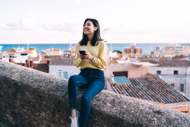 Cheerful Asian brunette female in casual clothes with toothy smile sitting on stone parapet and browsing smartphone while looking away - Fotoğraf, Görsel