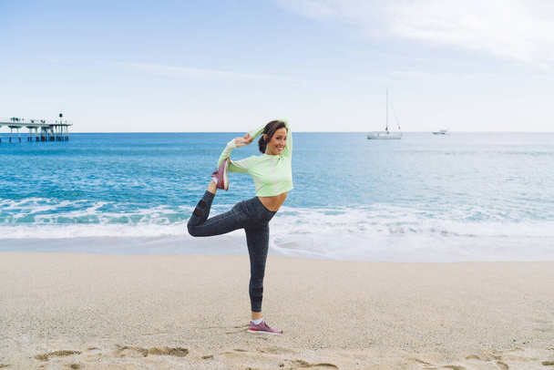 Full body charming adult female in sportswear standing in balance posture while doing yoga exercises on seashore with sea waves running on background - Foto, imagen