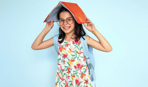 Happy student girl putting and holding academic book on her head and looking at the camera, wearing colorful dress, eyeglasses, and backpack posing on blue studio background. - Foto, Bild