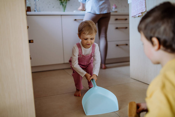 Un ragazzino e una ragazza che aiutano a pulire la casa usando padella e spazzola mentre spazzano via la sporcizia dal pavimento. - Foto, immagini