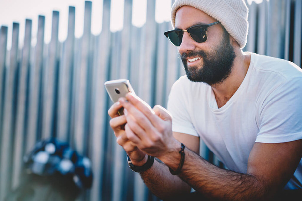 Middle Eastern male in trendy sunglasses and winter hat using smartphone gadget for browsing banking application and checking account details - Фото, зображення