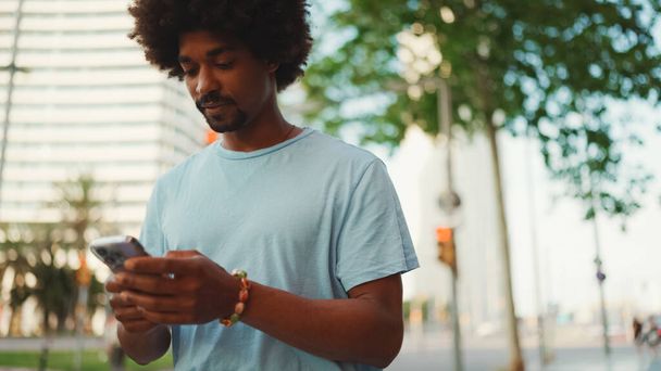 Closeup portrait of  young African American man in light blue t-shirt using his smartphone. Man looks at photos, videos in his mobile phone.  - Photo, Image