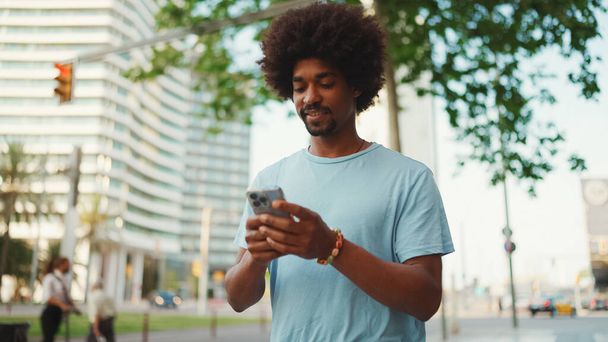 Closeup portrait of  young African American man in light blue t-shirt using his smartphone. Man looks at photos, videos in his mobile phone.  - Zdjęcie, obraz