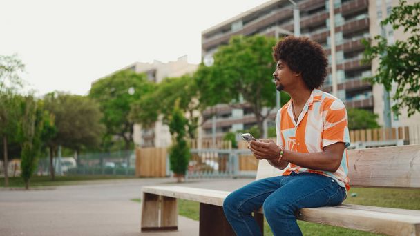 Close-up of young African American man wearing shirt sitting on park bench using his smartphone. Closeup of smiling african american man looking at photo video on mobile phone. Lifestyle concept - Photo, Image