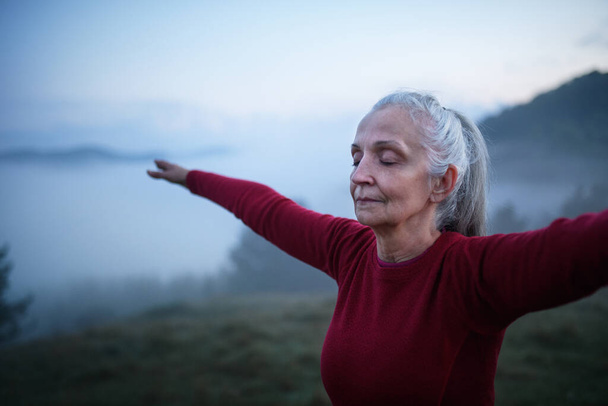 A senior woman doing breathing exercise in nature on early morning with fog and mountains in background. - Valokuva, kuva