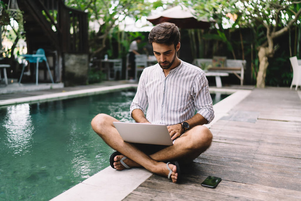 Young bearded male entrepreneur in summer shirt and flip flops working on laptop while sitting on floor near swimming pool in quiet backyard - Zdjęcie, obraz