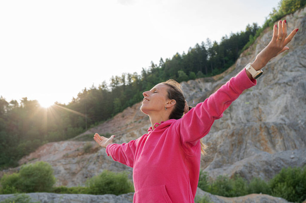 Γαλήνια νεαρή γυναίκα με ροζ πουλόβερ στέκεται έξω την αυγή με τα χέρια της ορθάνοιχτα, γιορτάζοντας τη ζωή και την ελευθερία. - Φωτογραφία, εικόνα