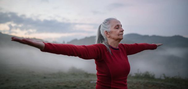 A senior woman doing breathing exercise in nature on early morning with fog and mountains in background. - Fotoğraf, Görsel