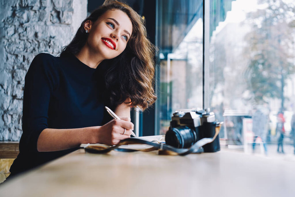 Positive female freelancer photography with red lips looking away and taking note while sitting at table with notebook and photo camera in modern workspace - Foto, afbeelding