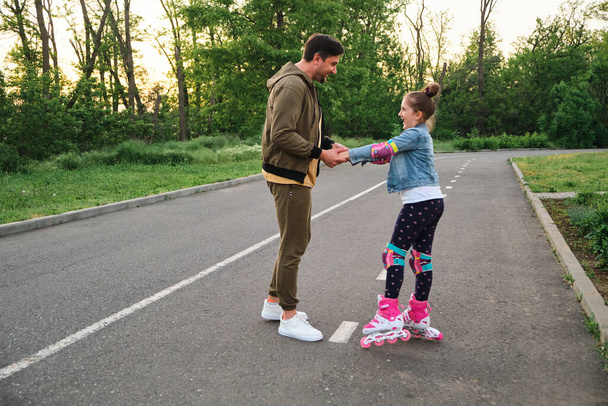 Vader leert zijn dochter schaatsen op rolschaatsen. Gelukkige jongen die schaatst. Familie brengt tijd samen door. Zonnige zomerdag in de buitenwijk straat - Foto, afbeelding