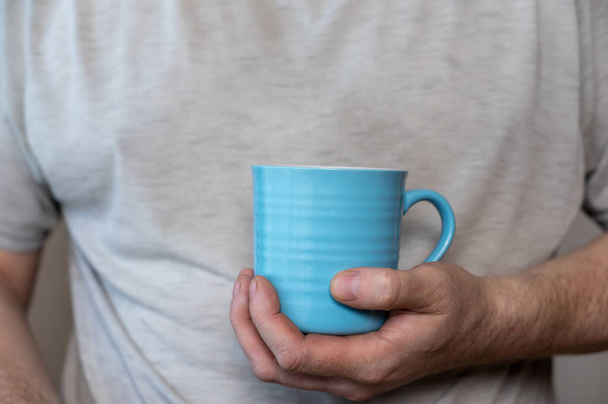 A middle-aged man is holding a blue mug. A ceramic cup with a handle for hot drinks in a man's hand. Middle section of a mature man in a gray T-shirt. Inside the room. Selective Focus. - Photo, Image