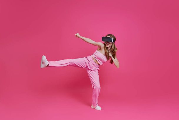 Playful young woman in virtual reality headset imitating fight while standing against pink background - Fotó, kép
