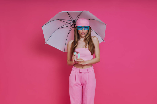 Attractive young woman in trendy clothing holding umbrella while standing against pink background - Zdjęcie, obraz