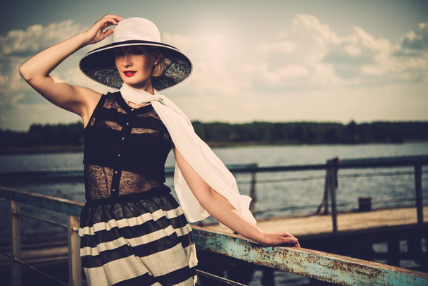 Beautiful woman wearing hat and white scarf sitting on old wooden pier  - Foto, Bild