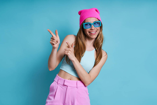 Attractive young woman in pink hat smiling and pointing away while standing against blue background - Photo, image