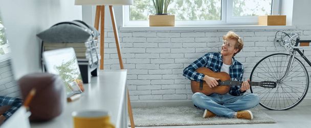 Happy young man playing acoustic guitar while sitting on the floor at home - Zdjęcie, obraz
