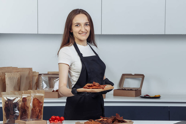 Meisje met vlees op een dienblad. Hoge kwaliteit een meisje houdt vlees op een dienblad op een lichte achtergrond met een keuken in zwarte wanten - Foto, afbeelding