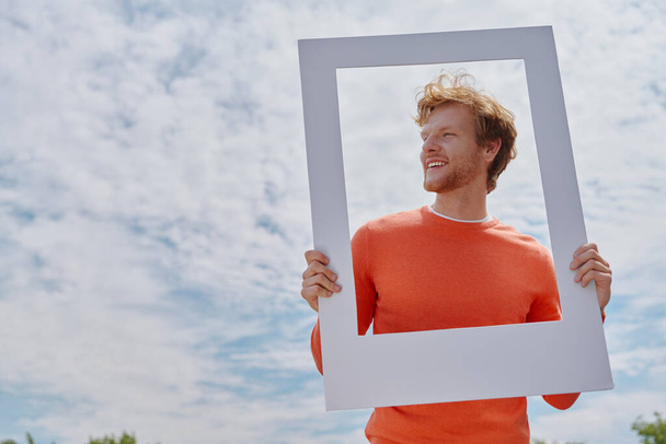 Playful young redhead man looking through a picture frame and smiling while standing outdoors - Fotoğraf, Görsel