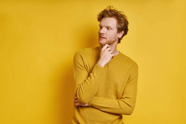 Handsome redhead man holding hand on chin while standing against yellow background - Фото, зображення