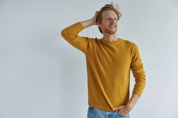 Cheerful young redhead man holding hand in hair while standing against wall - Fotografie, Obrázek