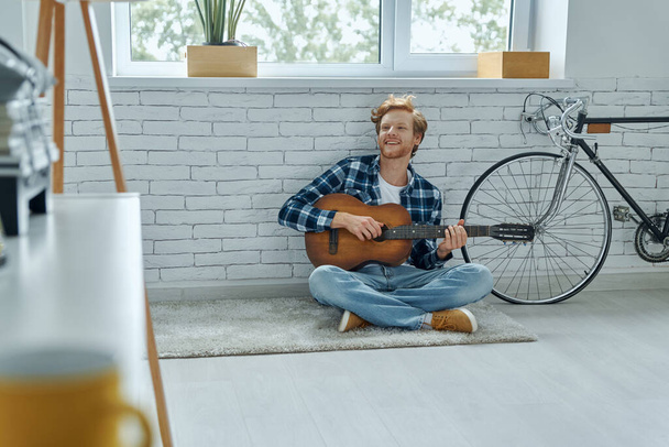 Evde yerde otururken akustik gitar çalan yakışıklı genç adam. - Fotoğraf, Görsel