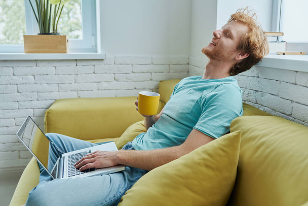 Relaxed redhead man using laptop and enjoying hot drink while sitting on the couch at home - Foto, Imagem