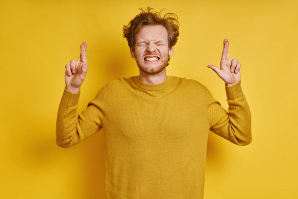 Young redhead man keeping fingers crossed while standing against yellow background - Foto, afbeelding