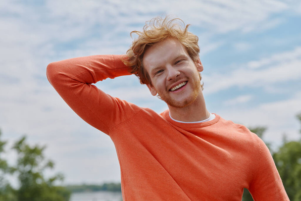 Relaxed young redhead man holding hand behind head and smiling while standing outdoors - Foto, Imagem