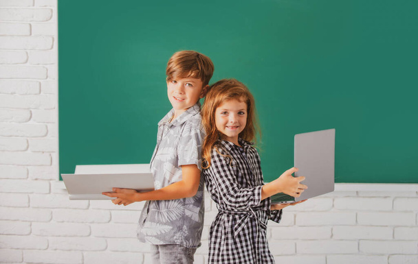 Muotokuva lasten poika ja tyttö kannettavan tietokoneen ja kirjan koulussa. Lapset luokassa liitutaulussa - Valokuva, kuva