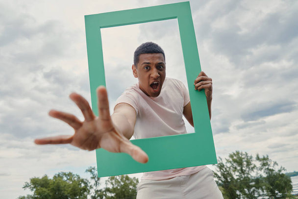 Playful mixed race man stretching out hand from picture frame with sky in the background - Fotoğraf, Görsel
