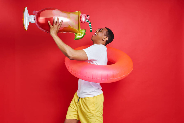 Playful young man carrying inflatable ring and cocktail while standing against red background - Photo, image