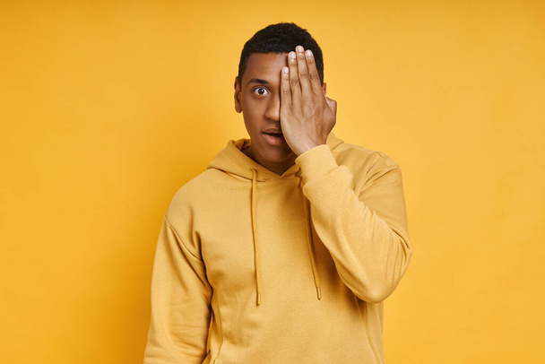 Shocked mixed race man covering half face with hand while standing against yellow background - Fotó, kép