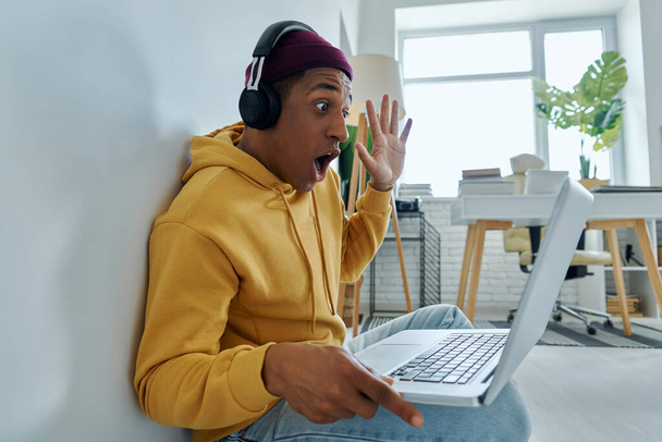 Excited man in headphones using laptop and gesturing while sitting on the floor at home - 写真・画像