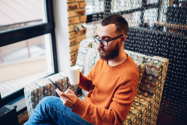 Young bearded hipster in trendy outfit and eyeglasses checking notifications on mobile phone and drinking hot beverage in cozy cafeteria - Photo, Image