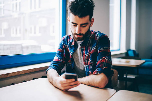 Bearded hipster guy dressed in casual wear messaging in online chat on modern smartphone using free 4G internet connection sitting at desktop in college.Student updating profile in social networks - Foto, Bild