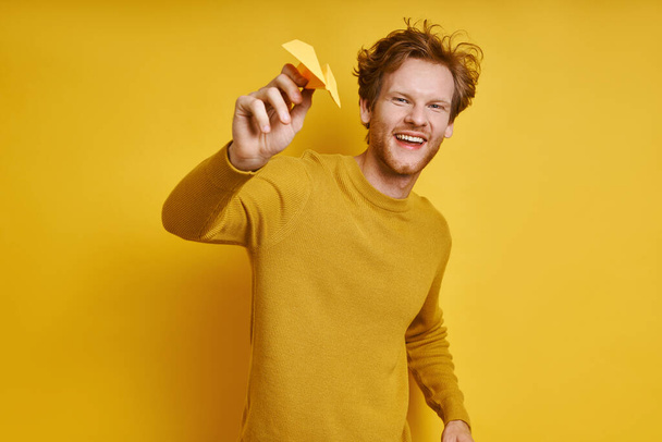 Ευτυχισμένος κοκκινομάλλης άνδρας κρατώντας χάρτινο αεροπλάνο και χαμογελώντας ενώ στέκεται απέναντι σε κίτρινο φόντο - Φωτογραφία, εικόνα