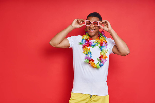 Happy young man in Hawaiian necklace standing against red background - Фото, зображення