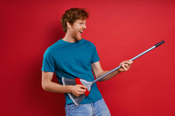 Playful young redhead man using broom like guitar while against red background - 写真・画像