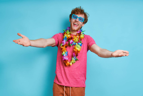Cheerful man in Hawaiian necklace gesturing welcome sign against blue background - Фото, изображение