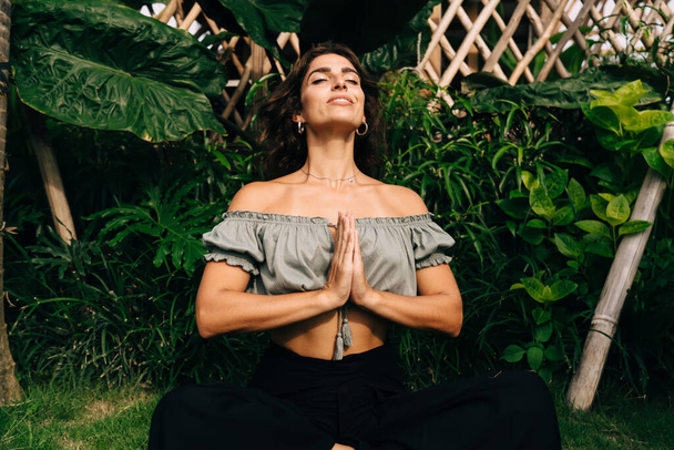 Happy young ethnic female traveler in trendy clothes doing yoga meditation with namaste gesture in Lotus pose during summer holidays in tropical country - Fotó, kép