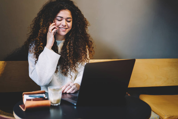 Fröhliche Hipster-Studentin mit lockigem Haar genießt Smartphone-Gespräche beim Laptop-Lernen in Coworking - Foto, Bild