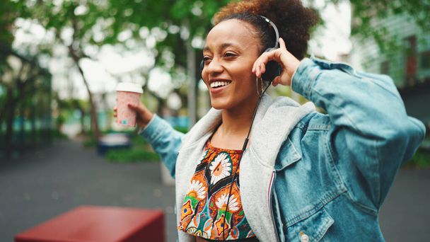 Smiling African girl with ponytail wearing denim jacket, in crop top with national pattern walking down the street listening to music on headphones and dancing - Fotó, kép