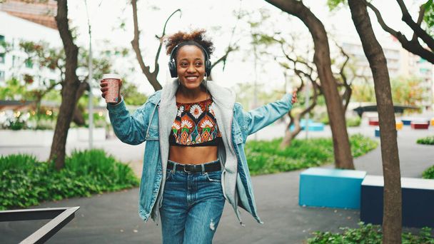 Smiling African girl with ponytail wearing denim jacket, in crop top with national pattern walking down the street listening to music on headphones and dancing - Photo, image