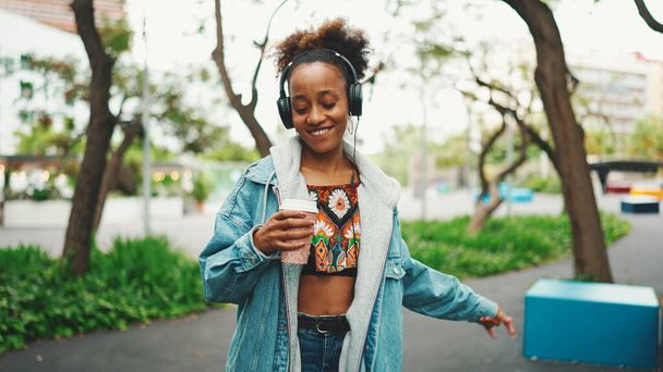Smiling African girl with ponytail wearing denim jacket, in crop top with national pattern walking down the street listening to music on headphones and dancing - 写真・画像