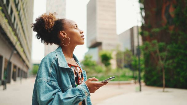 Close-up African girl wearing denim jacket, in crop top with national pattern goes on the street of modern city and holding smartphone looking at phone screen look for address with electronic map in city - Foto, Imagen