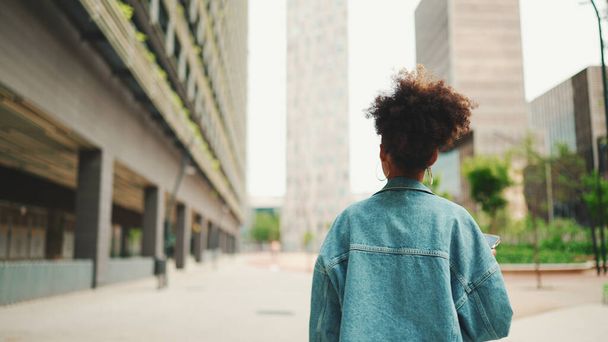 Close-up African girl wearing denim jacket, in crop top with national pattern goes on the street of modern city and holding smartphone looking at phone screen look for address with electronic map in city - 写真・画像