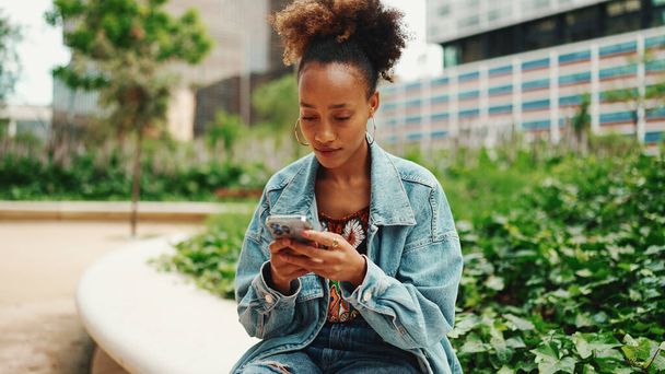 African girl with ponytail wearing denim jacket leaving voice message on mobile phone against modern city background.  - Fotografie, Obrázek