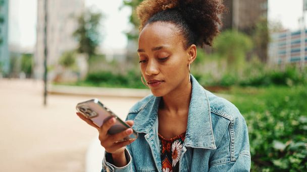 African girl with ponytail wearing denim jacket leaving voice message on mobile phone against modern city background.  - Foto, Bild
