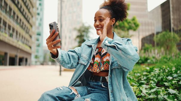 Closeup portret cute African girl with ponytail, wearing denim jacket makes stream, video call on smartphone in modern city background.  - Fotografie, Obrázek
